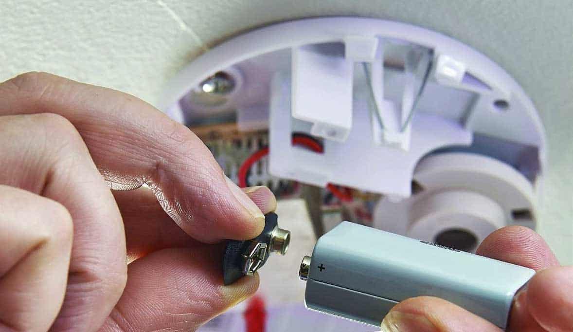 A person installing a battery into a carbon monoxide detector mounted on a ceiling.