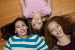 Three children lying on a wooden floor, smiling up at the camera.