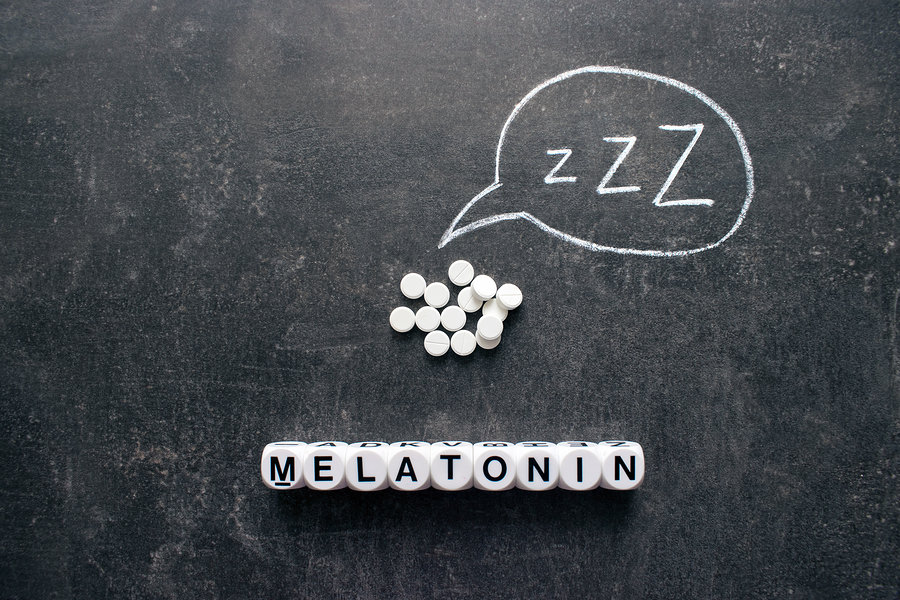Letter dice spelling 'Melatonin' on a dark surface, with small white tablets and a chalk speech bubble drawn above them containing 'ZZZ.'