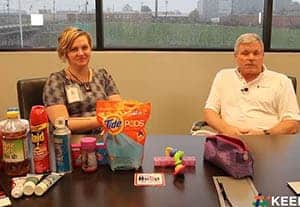 Two poison prevention experts seated at a table displaying common household items—including laundry detergent pods, cleaning products, medications, and personal care items—during a “Home Safety and Poison Prevention” press segment.