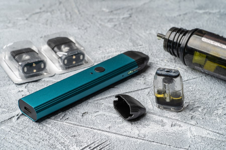 Vaping devices and accessories including a pod-style vape pen, replacement pods, and a larger tank-style vaporizer on a gray stone surface