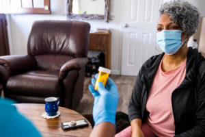 Healthcare worker wearing a mask and blue gloves handing a prescription bottle to an older adult woman wearing a mask, in a home setting