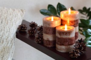 Three candles burning on a countertop besides some pinecones and a warm blanket.