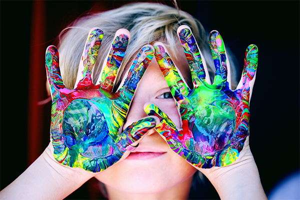 A young child holding up both hands covered in colorful paint, partially obscuring their smiling face.