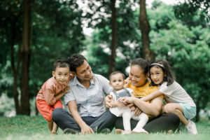 A family of five sitting together on grass in a park, with two parents and three young children smiling and embracing.