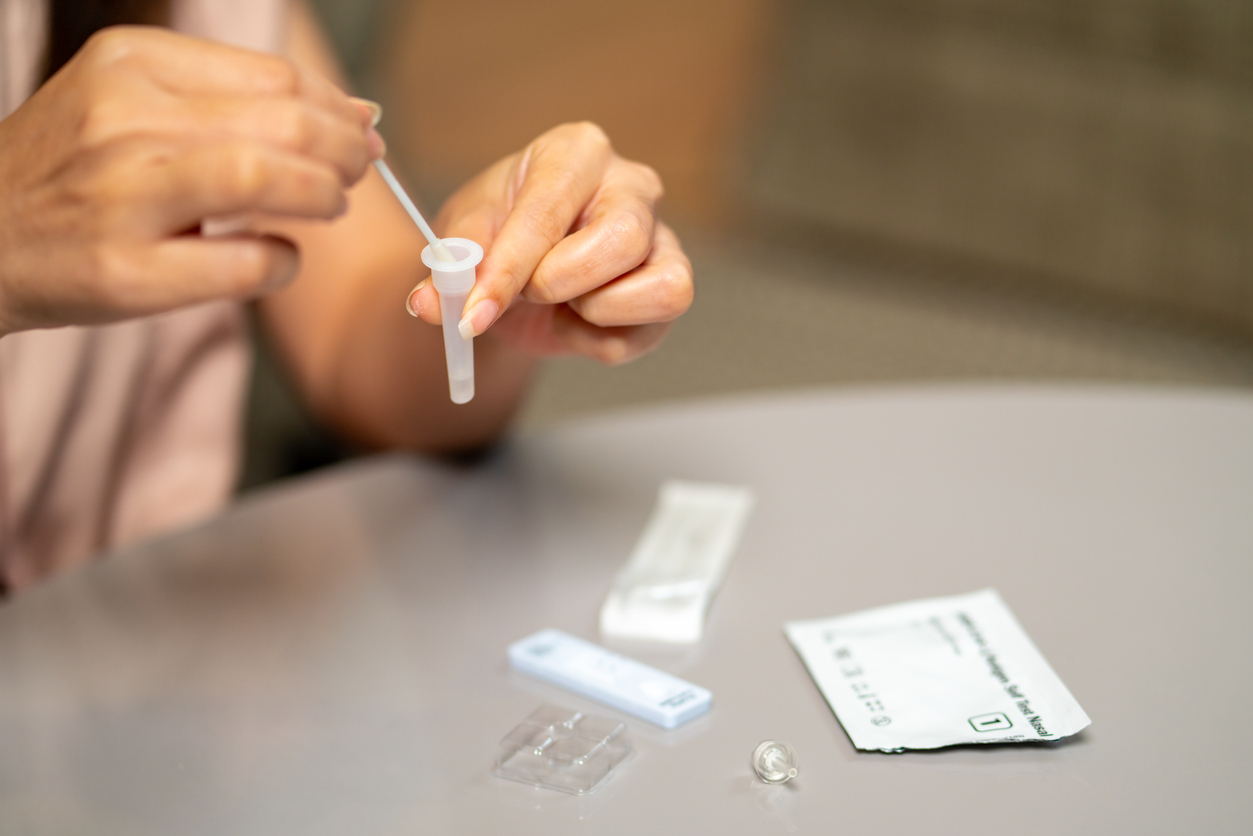 A person's hands using an at-home COVID-19 rapid test kit, with test components including a swab, vial, and test strip on a table.