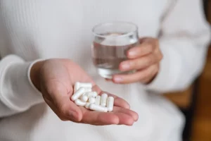 A woman holds a handful of white vitamin supplements in one hand. In the other, she holds a glass of water.