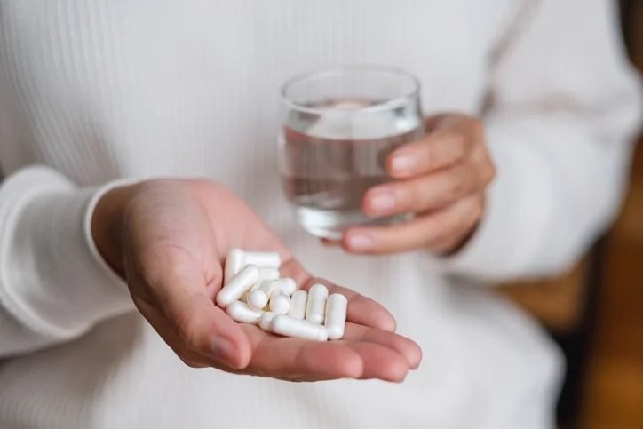 A woman holds a handful of white vitamin supplements in one hand. In the other, she holds a glass of water.
