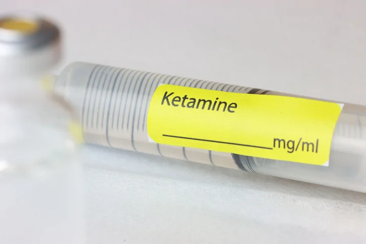 A plastic syringe set on a white table. It has a yellow label that reads "ketamine."