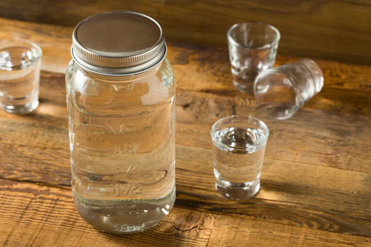 A mason jar of moonshine surrounded by 4 shot glasses.
